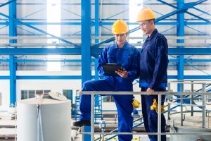 Two workers in large metal workshop or factory checking work standing on large machine